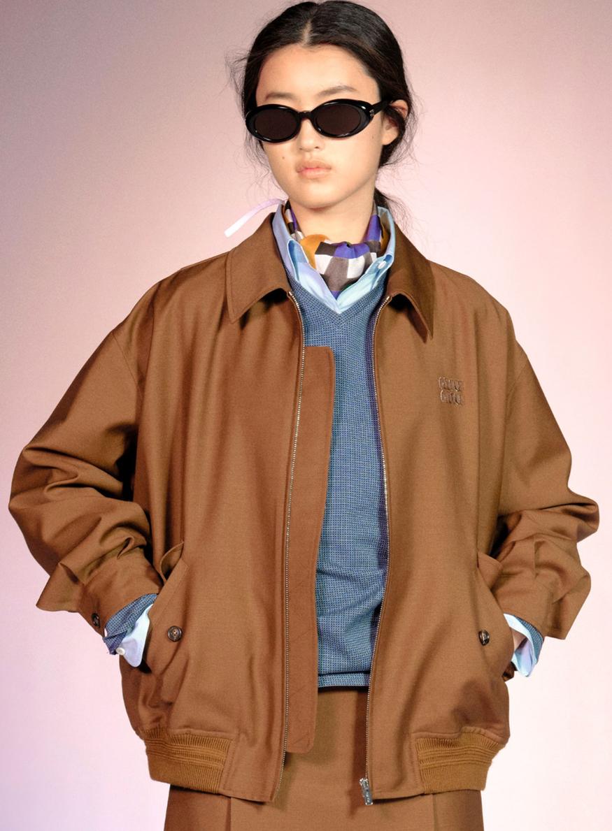 Silhouette Miu Miu collection 2025 — blouson bomber camel zippé porté sur pull col V bleu et chemise à carreaux, lunettes de soleil ovales noires
